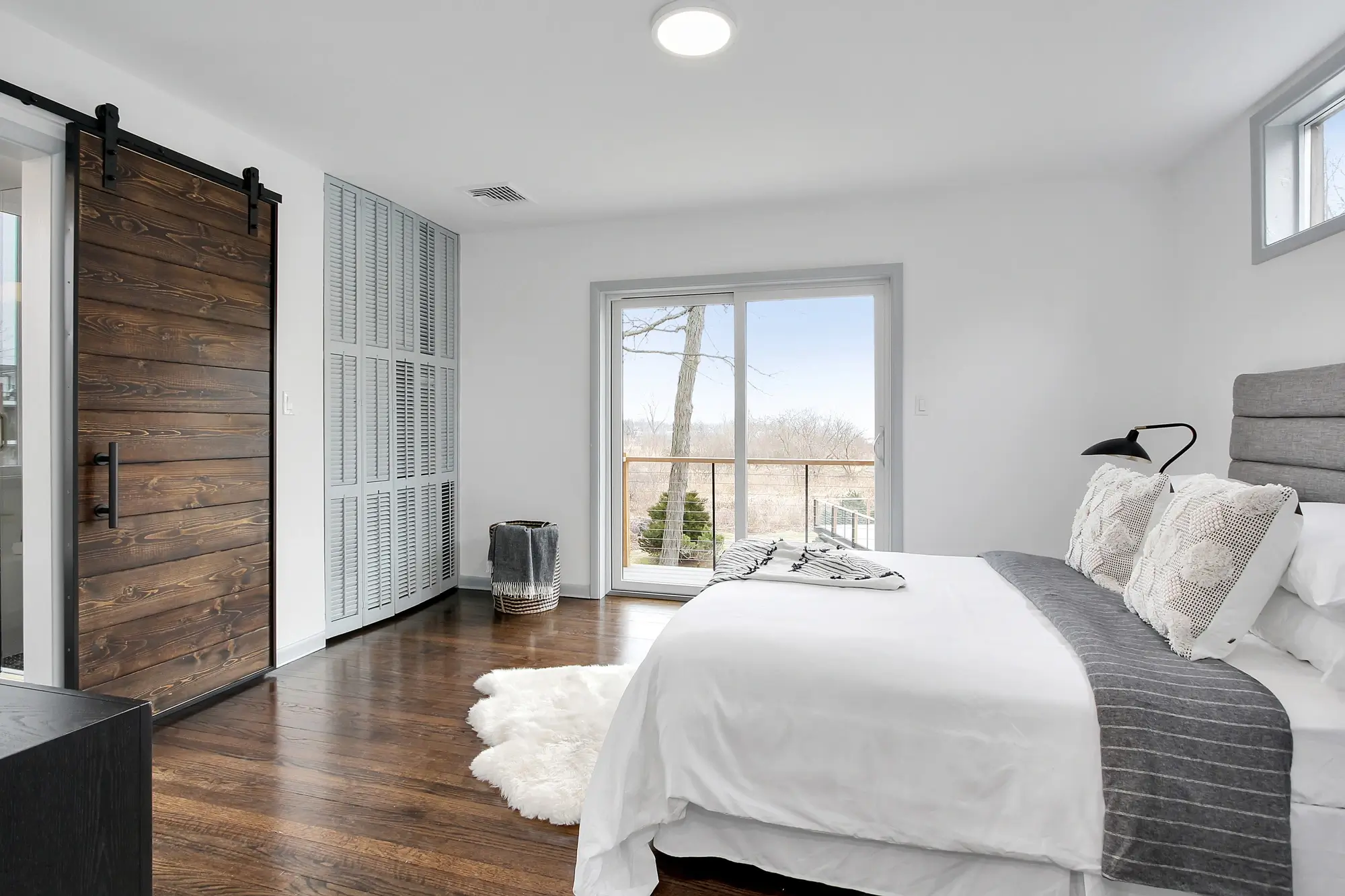 A room with a king size bed with pillows on top and comforter and a fluffy white rug on the floor and on the other side of the room there is a sliding door and outside the glass door you can see the beautiful scenery.
