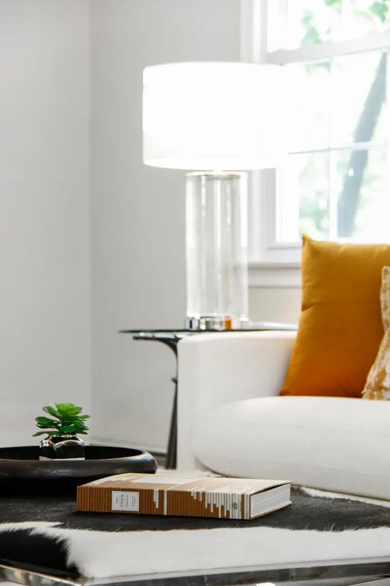 A white sofa with two pillows on it and next to it is a side table with a lamp on it and in front of the sofa is a center table with a book and a small plant on it.