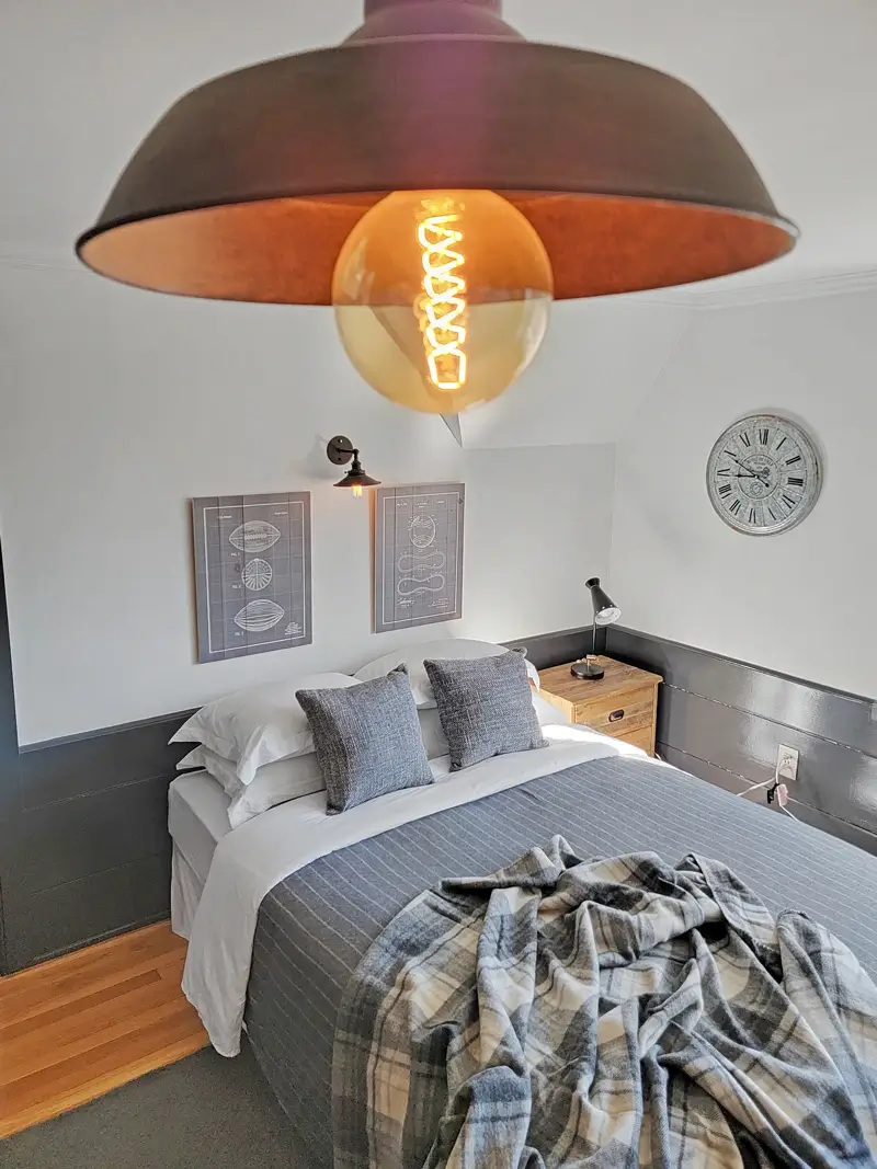 Double size bed with a pillows and gray comforter and blankets on it and one side table and two picture frames above the head board of the bed and on the right side of the wall a analog clock is hanging and on the ceiling there is a light with yellow bulb.