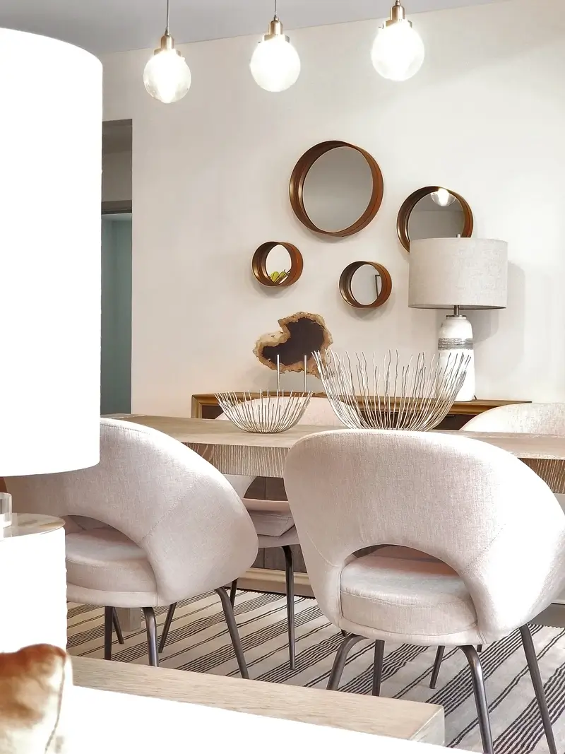 A beautiful dining area with four soft chairs and in the middle of the table there is a decorative display and behind the dining set there is a shelf and above it a lamp and on the wall there are round mirrors hanging.