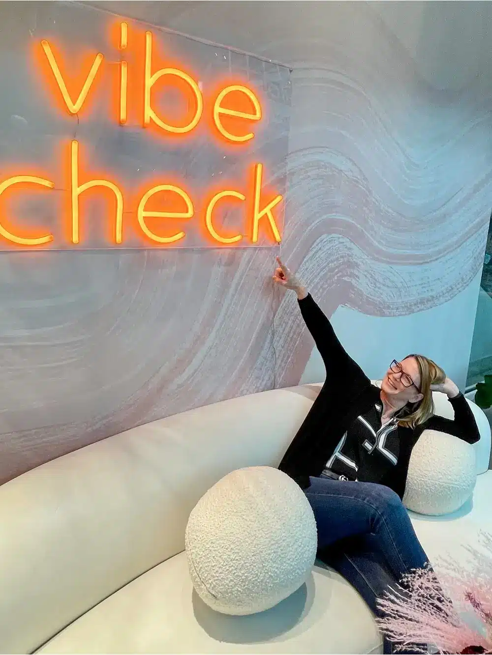 Picture of Birgit Anich - CEO seating on and white sofa with round shaped pillow on her both sides and she is smiling one of her hand pointing to the wall decorative display text "vibe check"