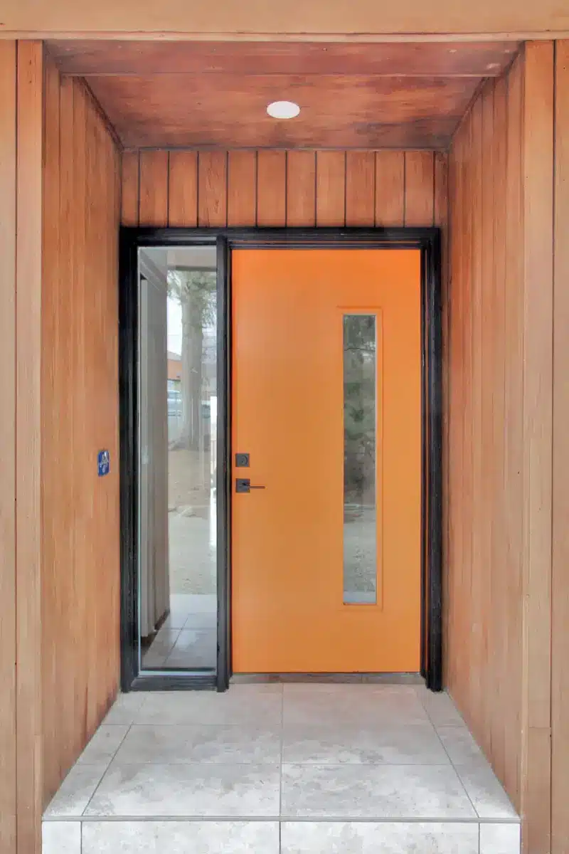 An orange door with a small glass window and another large glass window next to it and the wall is made of wood and the floor is paved.